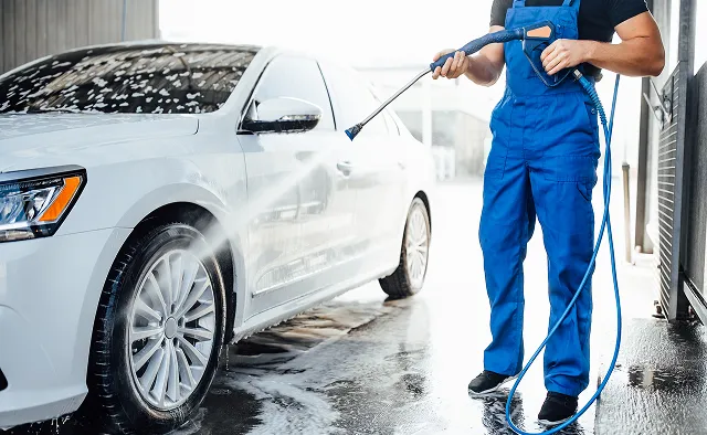 Man in blue overalls washing a white car with a pressure washer on a wet driveway.